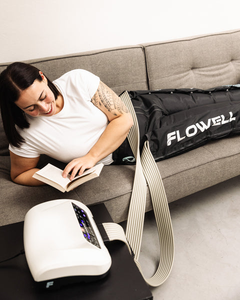 Flowell - Woman laying on a couch wearing her Flowell Home Deluxe Pants set and reading a book with the device's control unit by her side